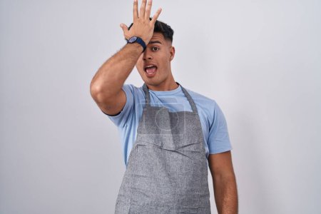 Photo for Hispanic young man wearing apron over white background surprised with hand on head for mistake, remember error. forgot, bad memory concept. - Royalty Free Image