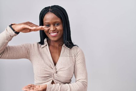 Foto de Mujer africana con trenzas de pie sobre fondo blanco haciendo gestos con las manos mostrando un signo de tamaño grande y grande, símbolo de medida. sonriendo mirando a la cámara. concepto de medición. - Imagen libre de derechos
