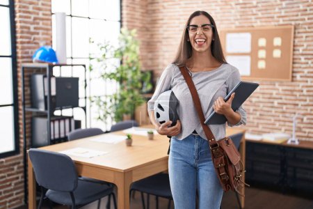 Photo for Young brunette woman working at the office holding bike helmet sticking tongue out happy with funny expression. - Royalty Free Image