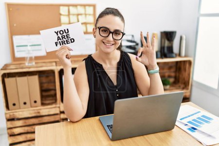 Photo for Young brunette woman holding you are fired banner at the office doing ok sign with fingers, smiling friendly gesturing excellent symbol - Royalty Free Image