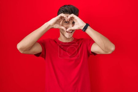Foto de Young hispanic man standing over red background doing heart shape with hand and fingers smiling looking through sign - Imagen libre de derechos