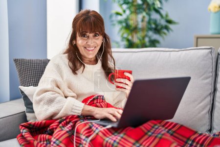 Photo for Middle age woman watching movie and drinking coffee sitting on sofa at home - Royalty Free Image