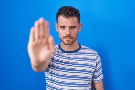 Téléchargez les photos : Jeune homme hispanique debout sur fond bleu faire arrêter chanter avec la paume de la main. expression d'avertissement avec geste négatif et sérieux sur le visage. - en image libre de droit