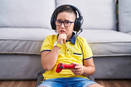 Foto de Joven niño hispano jugando videojuego sosteniendo controlador usando auriculares pensando preocupado por una pregunta, preocupado y nervioso con la mano en la barbilla - Imagen libre de derechos