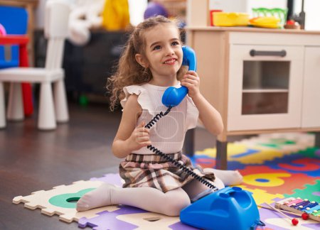 Photo for Adorable caucasian girl playing with telephone toy sitting on floor at classroom - Royalty Free Image