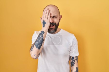 Foto de Joven hombre hispano con tatuajes de pie sobre fondo amarillo bostezando cansado cubriendo media cara, ojos y boca con la mano. cara duele en el dolor. - Imagen libre de derechos