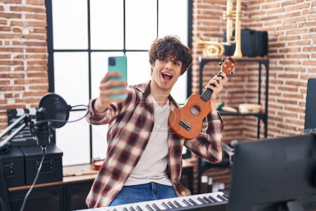 Foto de Young hispanic man musician making selfie by the smartphone holding ukelele at music studio - Imagen libre de derechos