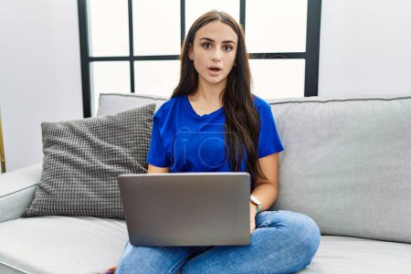Téléchargez les photos : Jeune femme brune utilisant un ordinateur portable à la maison effrayée et choquée par l'expression surprise, la peur et le visage excité. - en image libre de droit