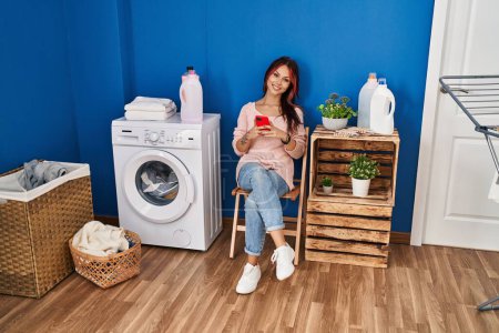 Foto de Mujer joven caucásica usando teléfono inteligente esperando a la lavadora en la sala de lavandería - Imagen libre de derechos