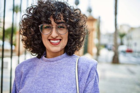 Téléchargez les photos : Jeune femme du Moyen-Orient souriant confiant portant des lunettes à la rue - en image libre de droit