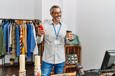 Téléchargez les photos : Moyen-âge homme aux cheveux gris boutique assistant en utilisant smartphone boire du café au magasin de vêtements - en image libre de droit