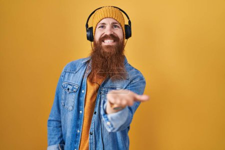 Foto de Hombre caucásico con barba larga escuchando música usando auriculares sonriendo alegre ofreciendo mano de palma dando asistencia y aceptación. - Imagen libre de derechos