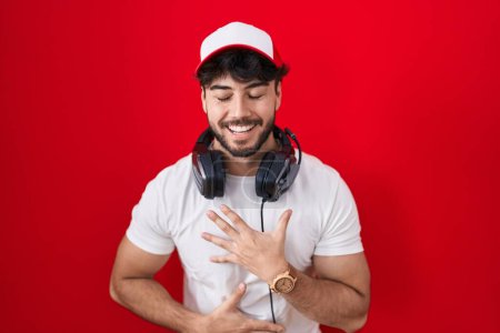 Foto de Hombre hispano con barba usando sombrero de gamer y auriculares sonriendo y riendo en voz alta porque divertido chiste loco con las manos en el cuerpo. - Imagen libre de derechos