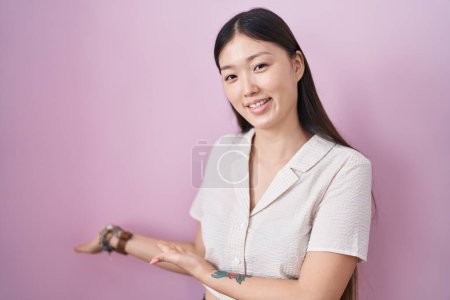 Photo for Chinese young woman standing over pink background inviting to enter smiling natural with open hand - Royalty Free Image