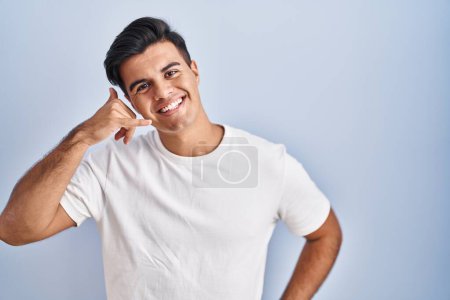 Téléchargez les photos : Homme hispanique debout sur fond bleu souriant faire geste de téléphone avec la main et les doigts comme parler au téléphone. communiquer les concepts. - en image libre de droit
