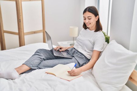 Téléchargez les photos : Jeune femme hispanique assise sur le lit à l'aide d'un ordinateur portable étudiant dans la chambre - en image libre de droit