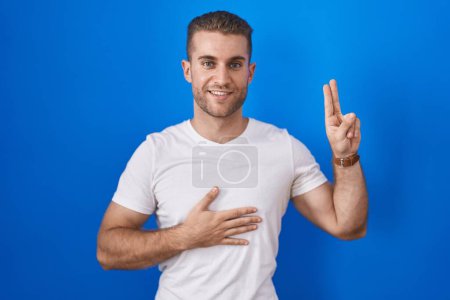 Téléchargez les photos : Young caucasian man standing over blue background smiling swearing with hand on chest and fingers up, making a loyalty promise oath - en image libre de droit