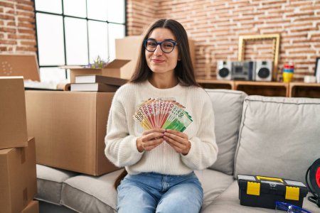 Photo for Young hispanic woman sitting on the sofa at new home holding money smiling looking to the side and staring away thinking. - Royalty Free Image