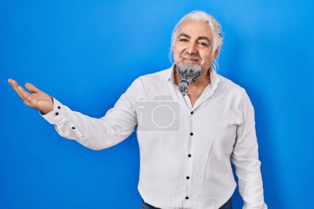 Foto de Hombre de mediana edad con el pelo gris de pie sobre fondo azul sonriendo alegre presentación y señalando con la palma de la mano mirando a la cámara. - Imagen libre de derechos