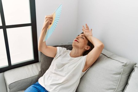 Photo for Middle age hispanic woman smiling sitting on the sofa using hand fan at home - Royalty Free Image