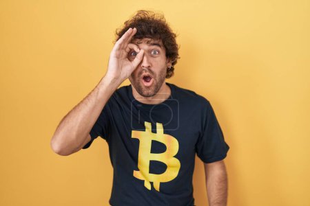 Photo for Hispanic young man wearing bitcoin t shirt doing ok gesture shocked with surprised face, eye looking through fingers. unbelieving expression. - Royalty Free Image