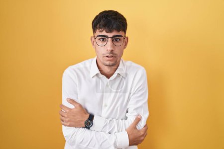 Foto de Young hispanic man standing over yellow background shaking and freezing for winter cold with sad and shock expression on face - Imagen libre de derechos