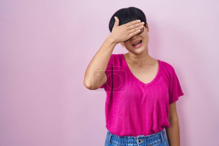 Foto de Joven mujer asiática con el pelo corto de pie sobre fondo rosa sonriendo y riendo con la mano en la cara cubriendo los ojos para sorpresa. concepto ciego. - Imagen libre de derechos