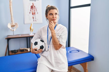 Foto de Young woman working at football therapy clinic serious face thinking about question with hand on chin, thoughtful about confusing idea - Imagen libre de derechos