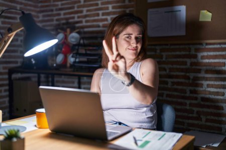 Téléchargez les photos : Femme brune travaillant au bureau la nuit souriant en regardant la caméra montrant les doigts faisant signe de victoire. numéro deux. - en image libre de droit