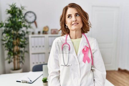 Foto de Médico de mediana edad mujer con cinta de cáncer rosa en uniforme mirando hacia un lado con sonrisa en la cara, expresión natural. riendo confiado. - Imagen libre de derechos