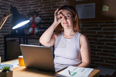 Photo for Brunette woman working at the office at night doing ok gesture with hand smiling, eye looking through fingers with happy face. - Royalty Free Image