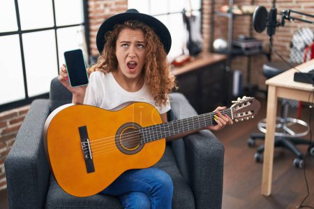 Photo for Young caucasian woman playing classic guitar at music studio holding smartphone angry and mad screaming frustrated and furious, shouting with anger looking up. - Royalty Free Image