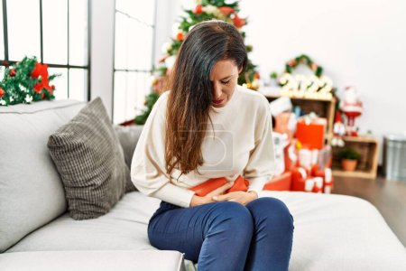 Photo for Middle age hispanic woman using hot water bag for period stomachache sitting by chrismas tree at home - Royalty Free Image