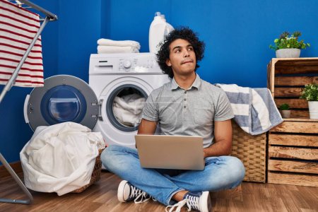 Téléchargez les photos : Jeune homme hispanique utilisant un ordinateur portable attendant la machine à laver à la buanderie - en image libre de droit