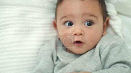 Téléchargez les photos : Adorable bébé hispanique couché sur le lit avec une expression détendue dans la chambre - en image libre de droit