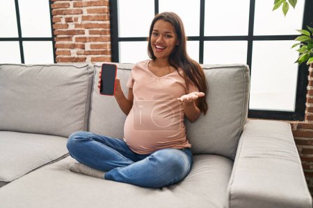 Téléchargez les photos : Jeune femme enceinte tenant smartphone montrant écran célébrant la réalisation avec sourire heureux et expression gagnante avec la main levée - en image libre de droit