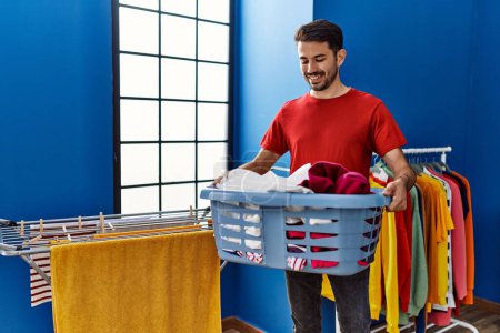 Photo for Young hispanic man smiling confident holding basket with clothes at laundry room - Royalty Free Image