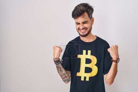 Foto de Young hispanic man with tattoos wearing bitcoin t shirt very happy and excited doing winner gesture with arms raised, smiling and screaming for success. celebration concept. - Imagen libre de derechos
