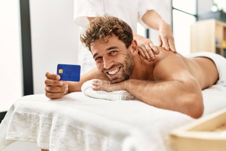 Photo for Young hispanic man having back massage holding credit card at beauty center - Royalty Free Image