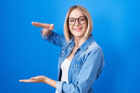 Téléchargez les photos : Jeune femme caucasienne debout sur fond bleu geste avec les mains montrant grand et grand signe de taille, symbole de mesure. souriant en regardant la caméra. concept de mesure. - en image libre de droit