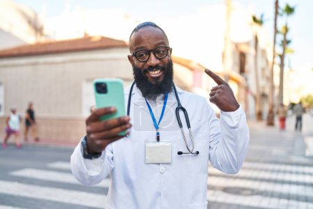 Photo for African american man wearing doctor uniform doing video call with smartphone smiling happy pointing with hand and finger - Royalty Free Image