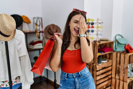 Photo for Young brunette woman holding shopping bags at retail shop covering one eye with hand, confident smile on face and surprise emotion. - Royalty Free Image
