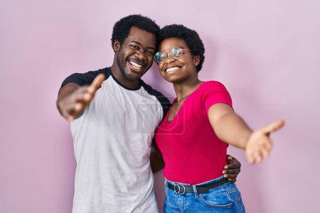 Téléchargez les photos : Jeune couple afro-américain debout sur fond rose regardant la caméra souriant à bras ouverts pour un câlin. expression joyeuse embrassant le bonheur. - en image libre de droit