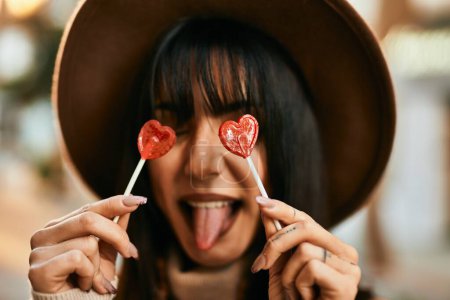 Photo for Brunette woman wearing winter hat being funny holding lollipops covering eyes outdoors at the city - Royalty Free Image