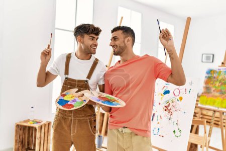 Foto de Dos hombres hispanos en pareja sonriendo confiados sosteniendo pinceles y paleta en el estudio de arte - Imagen libre de derechos