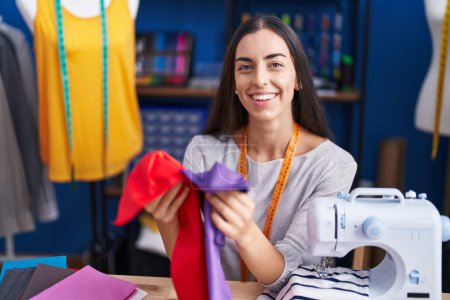 Foto de Joven hermosa mujer hispana sastre sonriendo confiado sosteniendo paños en la fábrica de ropa - Imagen libre de derechos