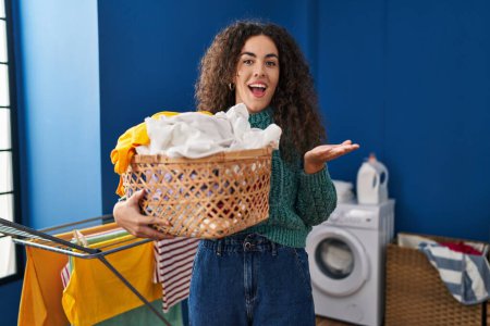 Photo for Young hispanic woman holding laundry basket celebrating achievement with happy smile and winner expression with raised hand - Royalty Free Image