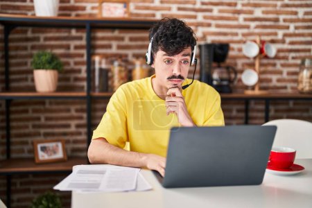 Photo for Hispanic man wearing call center agent headset working from home serious face thinking about question with hand on chin, thoughtful about confusing idea - Royalty Free Image