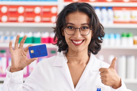 Photo for Young hispanic woman working at pharmacy drugstore holding credit card smiling happy and positive, thumb up doing excellent and approval sign - Royalty Free Image