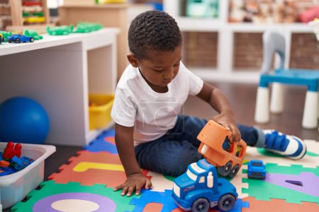 Téléchargez les photos : Africain américain garçon jouer avec des voitures et camion jouet assis sur le sol à la maternelle - en image libre de droit